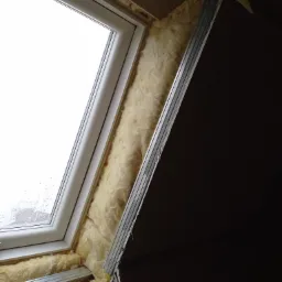 Okno dachowe podczas montażu, ocieplone wełną mineralną i zabezpieczone metalowym profilem w konstrukcji poddasza.