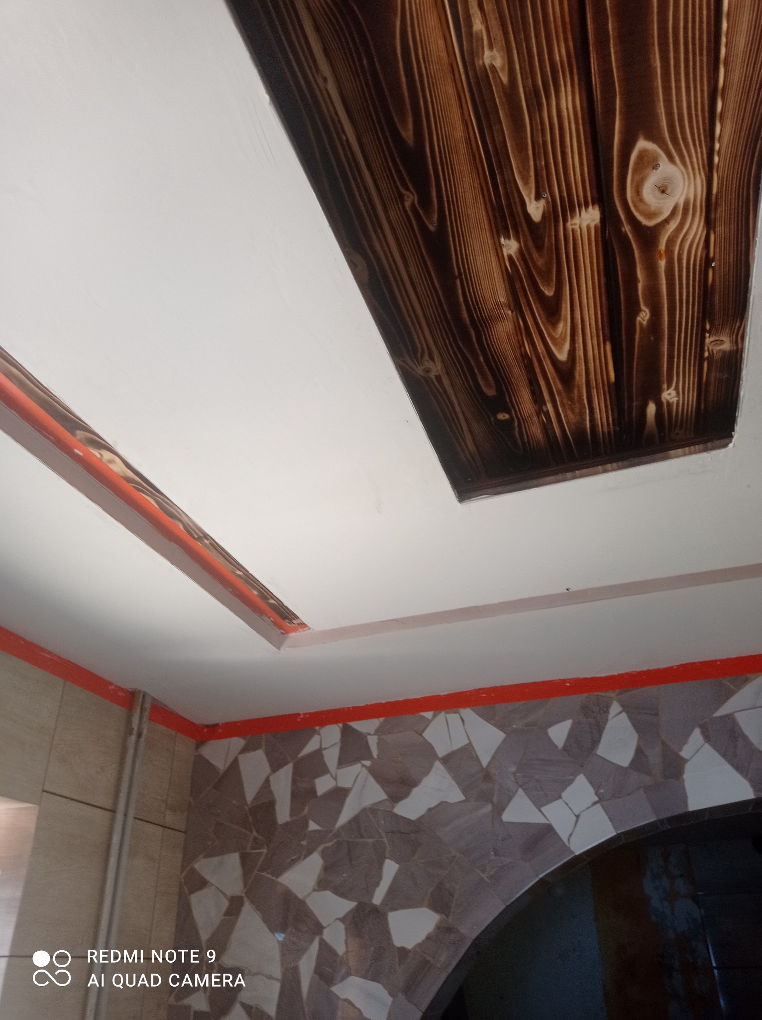 Fragment sufitu z wbudowanym, ciemnym, drewnianym panelem o wyraźnym usłojeniu i białymi płytami karton gips, wykończony pomarańczową listwą.