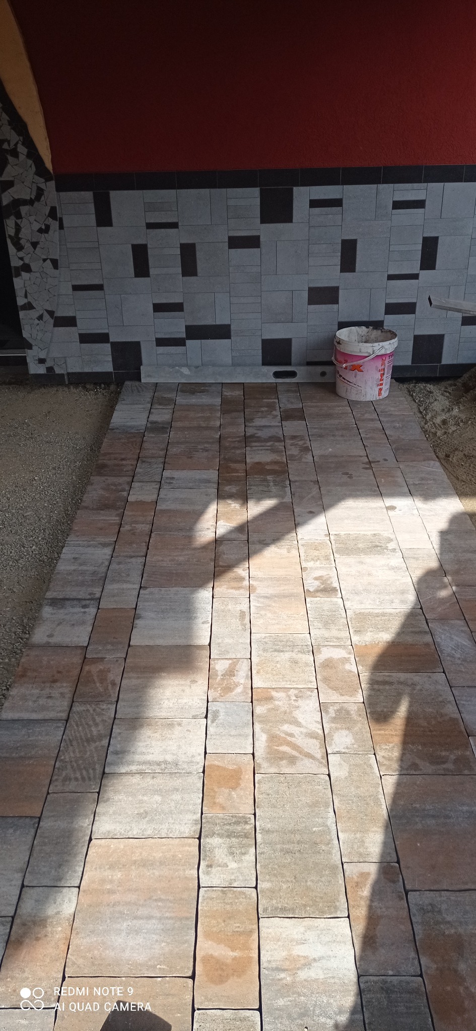 Ułożona kostka brukowa w odcieniach szarości i brązu prowadząca do ściany z dekoracyjną okładziną, obok stoi wiadro z resztkami zaprawy.