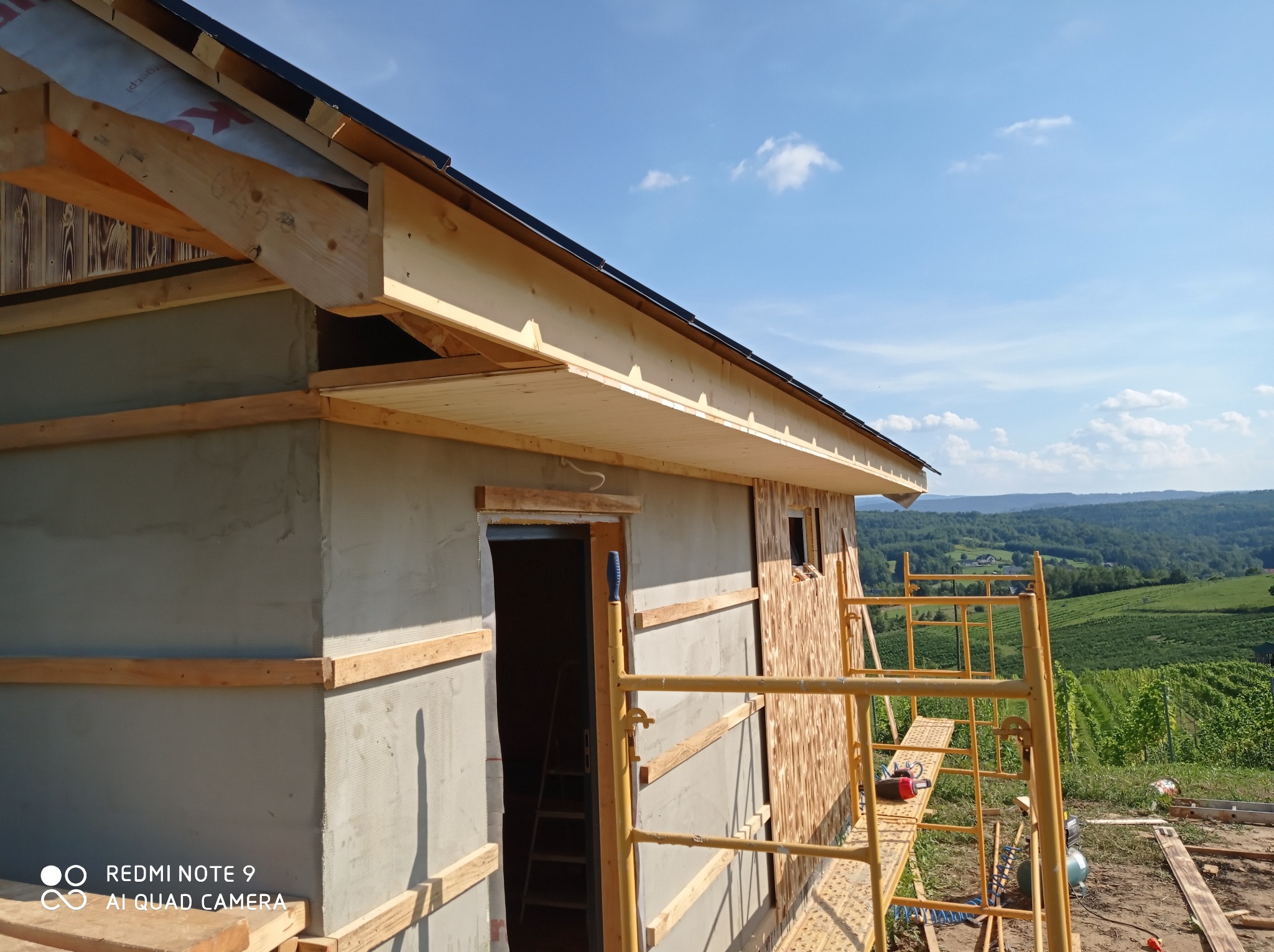 Budowa domu w trakcie ocieplania ścian zewnętrznych, widoczne rusztowanie i drewniana konstrukcja dachu na tle krajobrazu.