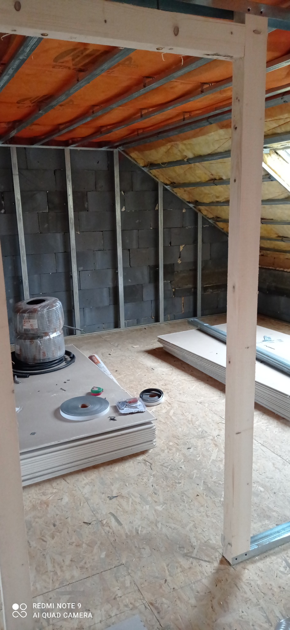 Poddasze w trakcie adaptacji: widoczne ocieplenie, metalowy stelaż, stos płyt kartonowo-gipsowych, rolka taśmy i narzędzia na podłodze OSB, drewniany słupek konstrukcyjny.