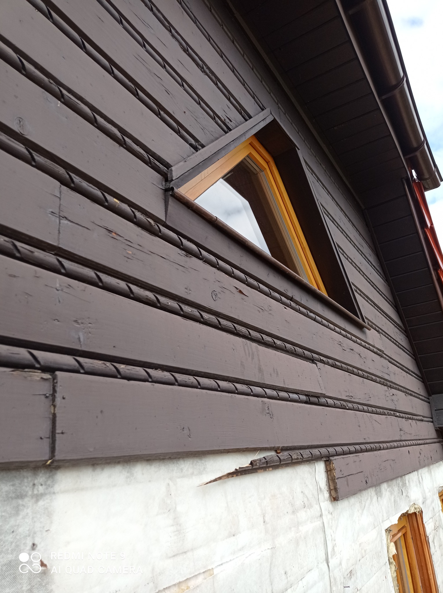 Fragment elewacji drewnianego domu z trójkątnym oknem, brązowe deski z dekoracyjnymi listwami, ocieplenie na dolnej części ściany.