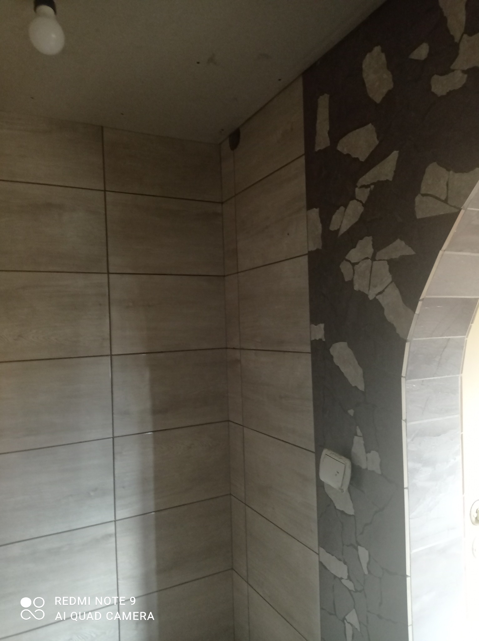 Wnętrze po remoncie: ściana wyłożona jasnymi, drewnopodobnymi płytkami ceramicznymi, sąsiadująca z fragmentem ściany z ciemnoszarą mozaiką z jasnymi elementami w kształcie nieregularnych odłamków...