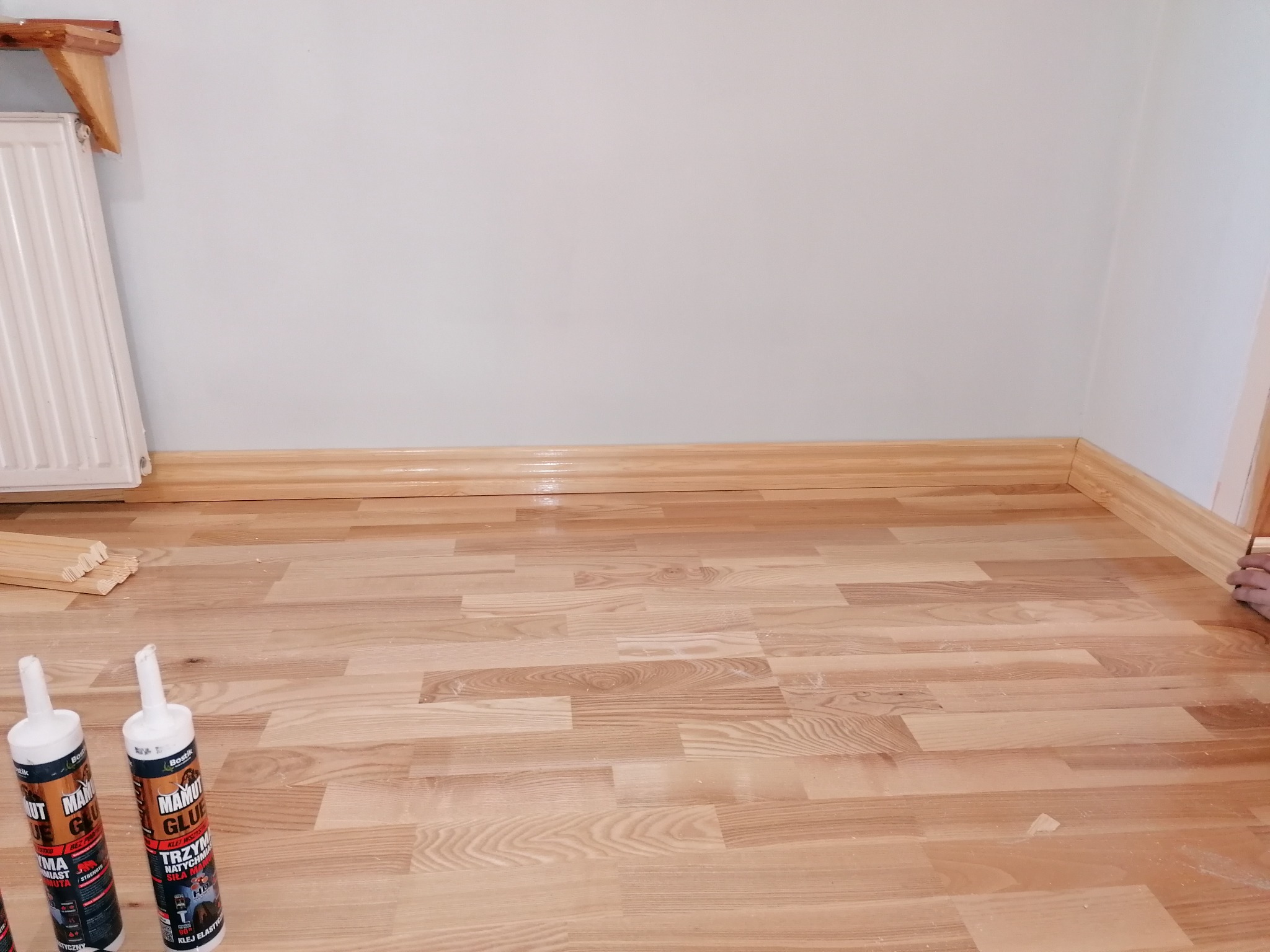 Jasne panele podłogowe ułożone w pokoju, widoczne listwy przypodłogowe i tuby kleju montażowego w rogu pomieszczenia.