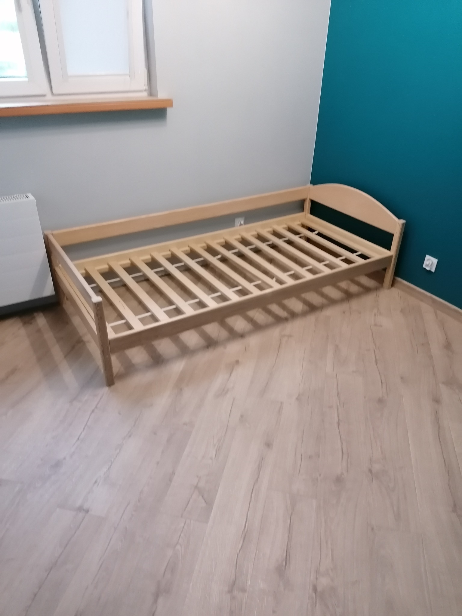 Surowa drewniana rama łóżka dziecięcego z listewkami, ustawiona na podłodze wyłożonej panelami imitującymi jasne drewno, na tle ściany pomalowanej na szaro i turkusowo.