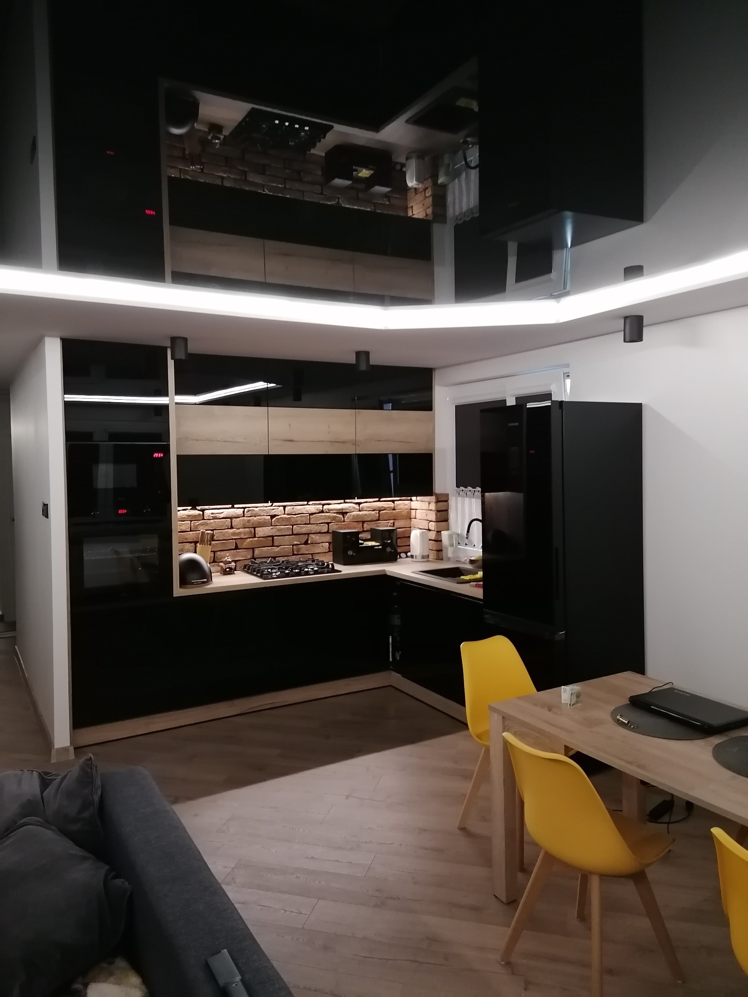 Nowoczesna kuchnia z czarnymi błyszczącymi frontami szafek, ceglaną ścianą nad blatem, oświetleniem LED i odblaskowym sufitem, widziana z perspektywy salonu z szarą sofą.