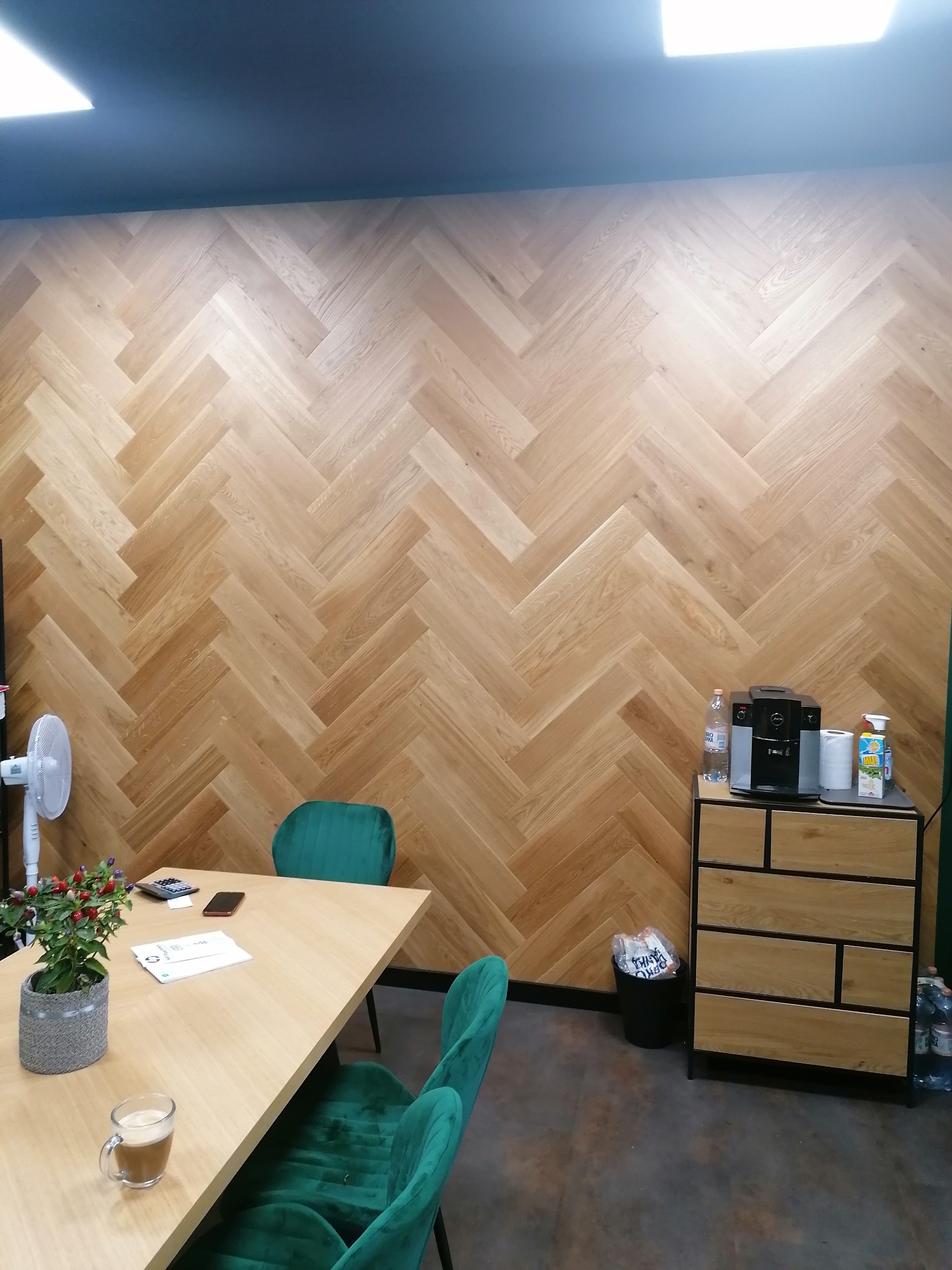 Ściana wykończona panelami ułożonymi w jodełkę, widoczny fragment biura z drewnianym biurkiem, zielonymi krzesłami i szafką z ekspresem do kawy.