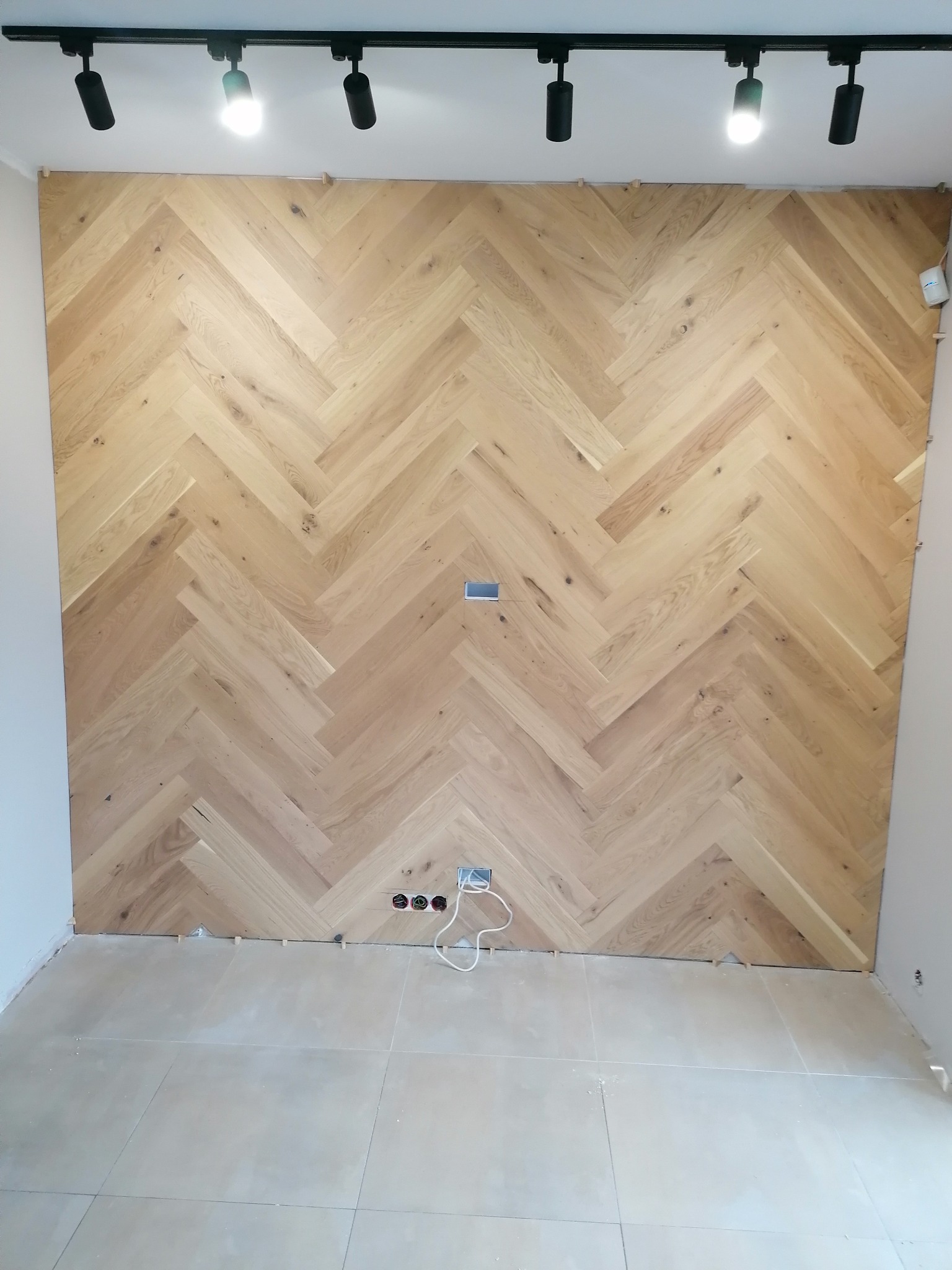 Ściana wyłożona parkietem w jodełkę, widoczne listewki dystansowe przy podłodze, gniazdka elektryczne i oświetlenie sufitowe punktowe.