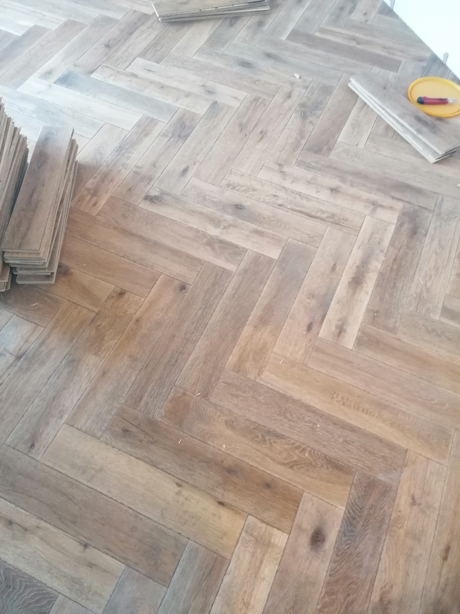 Ułożony parkiet w jodełkę z jasnego drewna, obok leżą nieużyte deski i nożyk w żółtym pudełku.