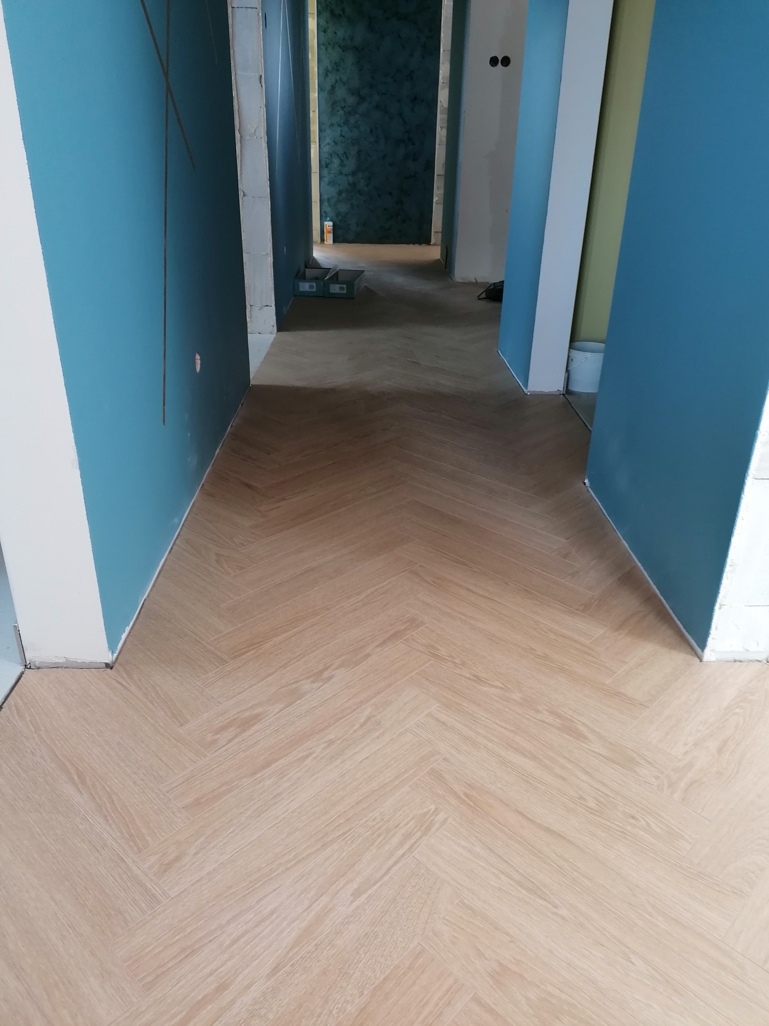 Jasna podłoga ułożona w jodełkę w trakcie remontu, widoczne świeżo pomalowane ściany na niebiesko i biało, surowy stan wykończenia w przejściu do kolejnych pomieszczeń.