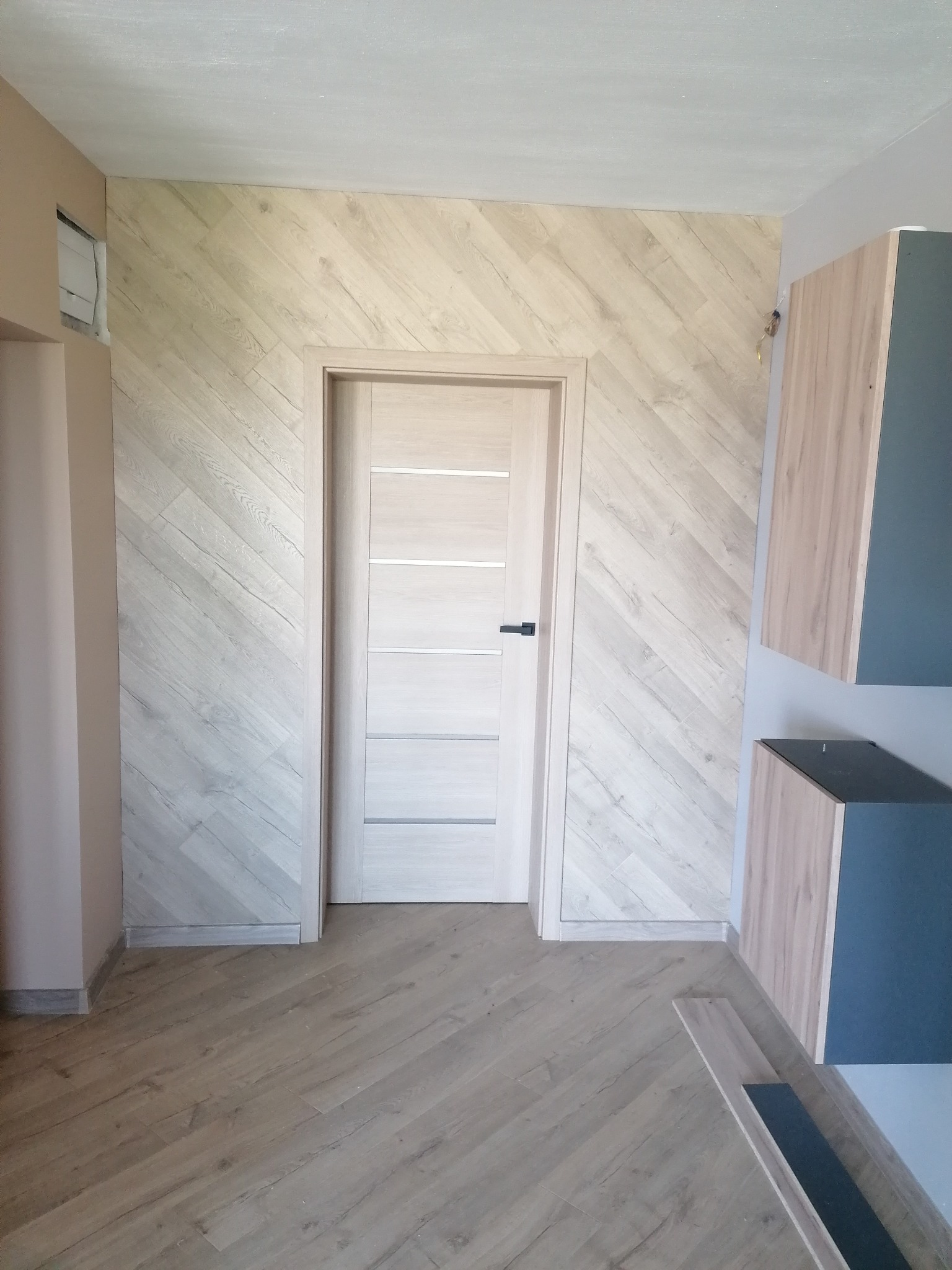 Wnętrze z panelami ułożonymi w jodełkę na ścianie i podłodze, jasne drzwi z poziomymi panelami i czarną klamką, fragment zabudowy kuchennej w tle.