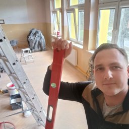 KOBER - Pracownik z poziomicą w dłoni, gotowy do malowania ścian w Świdnicy. W tle drabina, okna i przygotowane narzędzia malarskie. Widoczne ślady po remoncie.