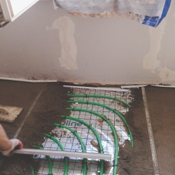 KOBER - Montaż ogrzewania podłogowego: zielone rurki na folii, częściowo zasypane piaskiem, wyrównywane łatą przez osobę w rękawicy. Widoczne okno i fragment ściany.