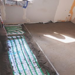 KOBER - Instalacja ogrzewania podłogowego z zielonymi rurami, częściowo zalana wylewką. Widoczny fragment pracownika w kombinezonie. W tle surowa ściana w trakcie remontu.