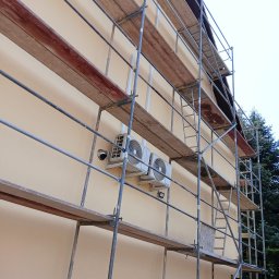 KOBER - Elewacja budynku w kolorze kremowym, z zamontowanymi dwoma klimatyzatorami i kamerą, otoczona rusztowaniem. Widok z dołu, częściowo widoczny dach i zieleń.