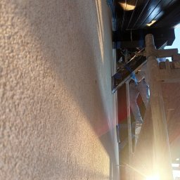 KOBER - Praca na rusztowaniu: aplikacja jasnego tynku strukturalnego na elewacji domu. Widoczne narzędzia i fragment osoby wykonującej prace. Silne światło słoneczne.