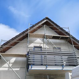 KOBER - Dom w trakcie prac elewacyjnych: rusztowanie, zabezpieczone okna, świeżo otynkowana ściana szczytowa pod brązowym dachem. Błękitne niebo w tle.