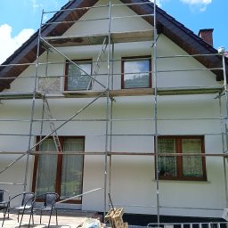 KOBER - Dom jednorodzinny w trakcie malowania elewacji, otoczony rusztowaniem. Widoczne materiały budowlane i narzędzia. Ujęcie w słoneczny dzień.