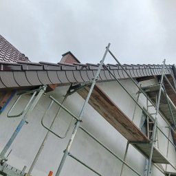 KOBER - Elewacja domu w trakcie prac: rusztowanie przy białej ścianie, widoczny fragment dachu z brązową dachówką. Ujęcie z dołu, pochmurne niebo w tle.