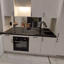 Marcin Budowlaniec - Nowoczesna kuchnia z białymi szafkami, czarnym blatem i marmurową podłogą; w tle widoczne odbicie osoby i drabiny.