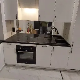 Nowoczesna kuchnia z białymi szafkami, czarnym blatem i marmurową podłogą; w tle widoczne odbicie osoby i drabiny.