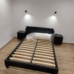 Marcin Budowlaniec - Rama łóżka z drewnianym stelażem, czarna tapicerka, dwie szafki nocne, materac w folii, oświetlenie punktowe na ścianie, panele podłogowe.
