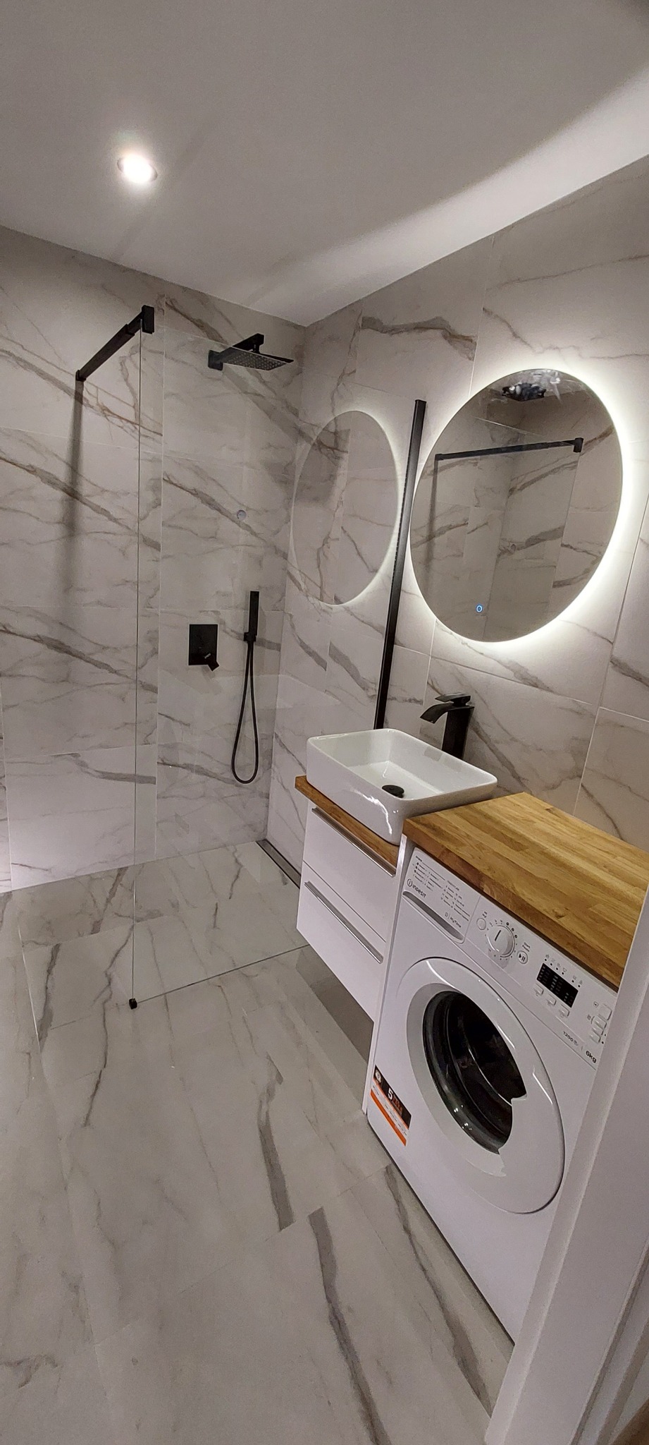 Nowoczesna łazienka z kabiną prysznicową typu walk-in, umywalką nablatową na szafce z drewnianym blatem, okrągłym lustrem z podświetleniem LED i pralką w zabudowie.