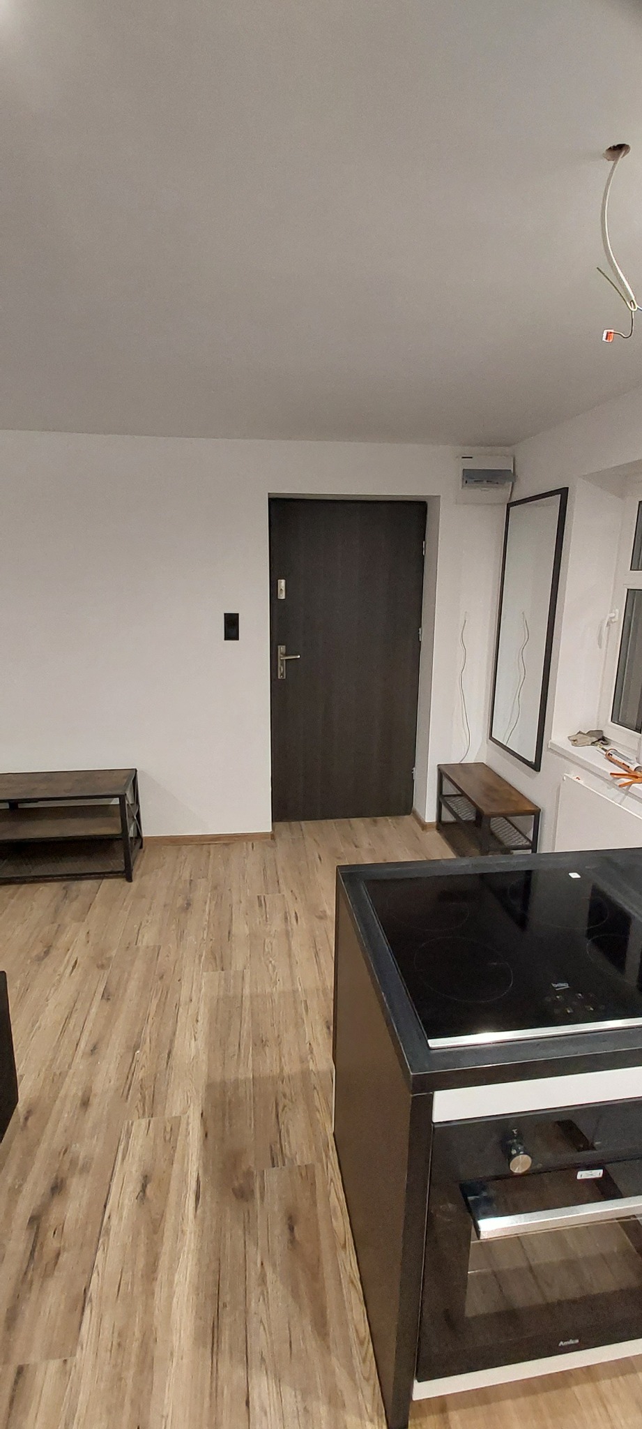 Wnętrze mieszkania po remoncie: widok na nowe panele podłogowe w odcieniach drewna, ciemne drzwi wejściowe, białe ściany, meble oraz fragment zabudowy kuchennej z piekarnikiem.