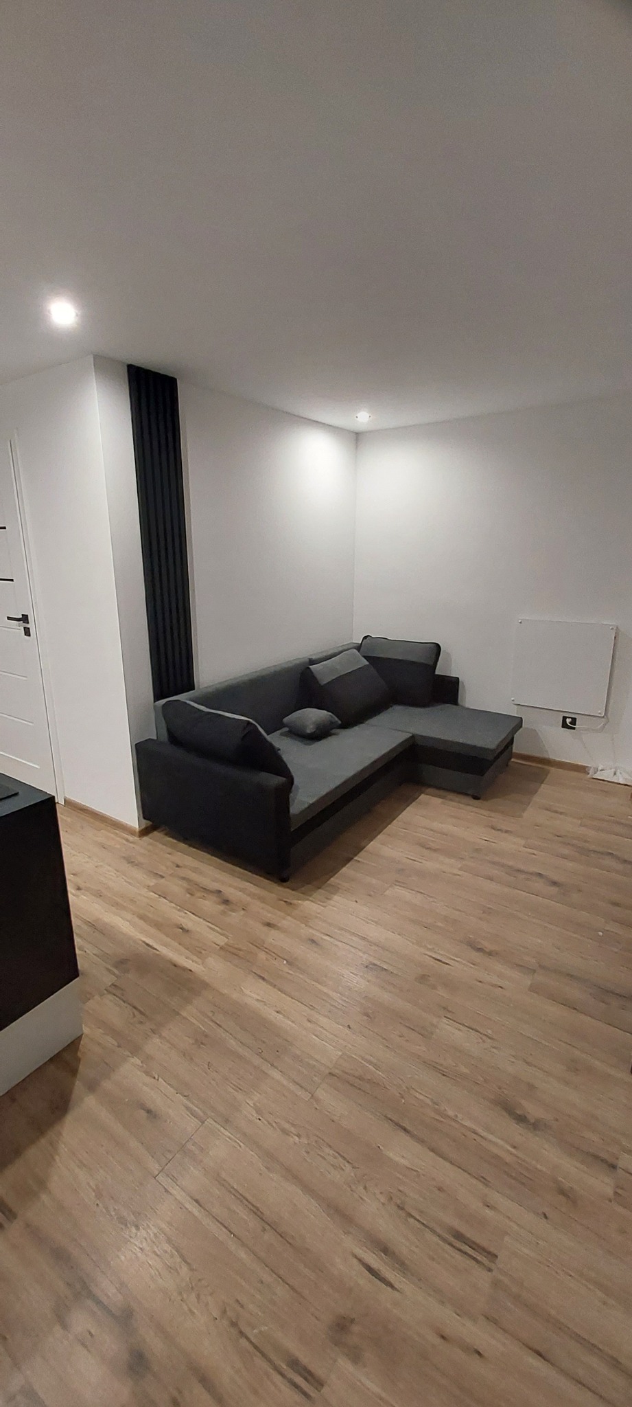 Nowoczesny narożnik w szaro-czarnych tonacjach na podłodze z paneli imitujących drewno, białe ściany z czarnym dekoracyjnym panelem pionowym i grzejnikiem elektrycznym.