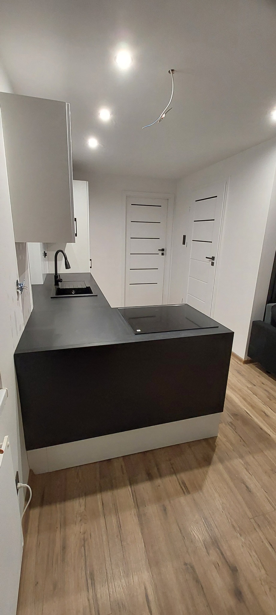 Nowoczesna kuchnia z czarnym blatem, wbudowaną płytą indukcyjną i minimalistycznym zlewem, widoczne dwie białe drzwi z czarnymi poziomymi paskami oraz fragment drewnopodobnej podłogi.