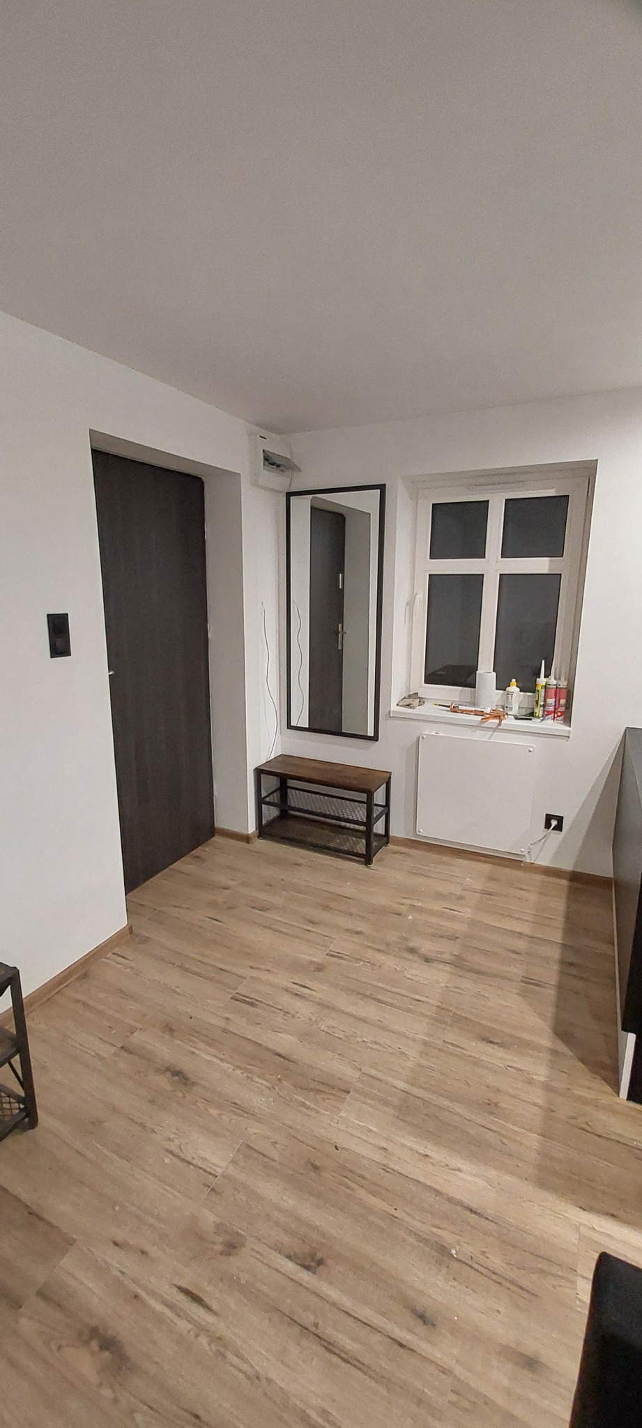 Wnętrze przedpokoju po remoncie z drewnopodobną podłogą, ciemnymi drzwiami i lustrem w czarnej ramie, widoczne ślady po pracach wykończeniowych na parapecie.