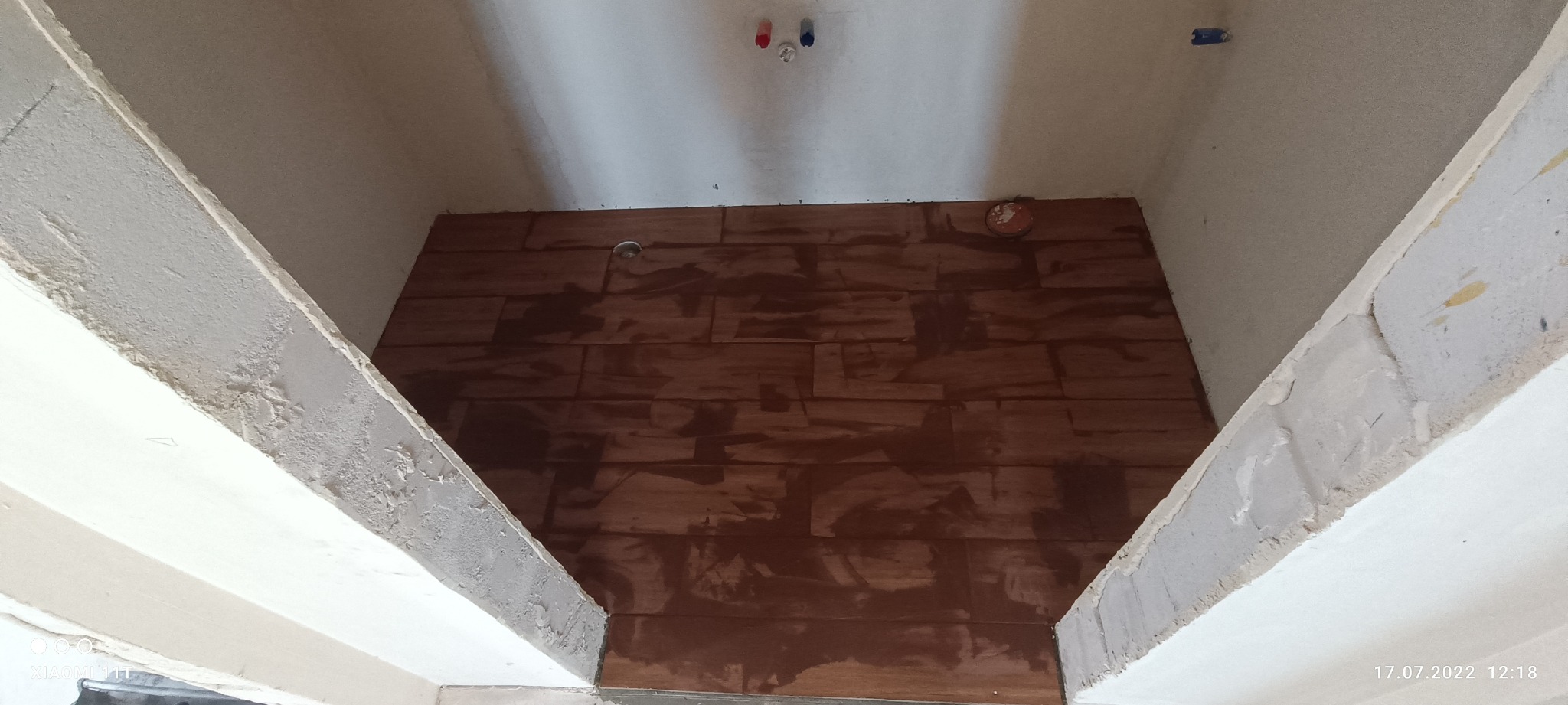 Wnęka z brązowymi płytkami imitującymi drewno, widoczne instalacje wodne i elektryczne na suficie, surowy stan wykończenia ścian i obramowania wnęki.