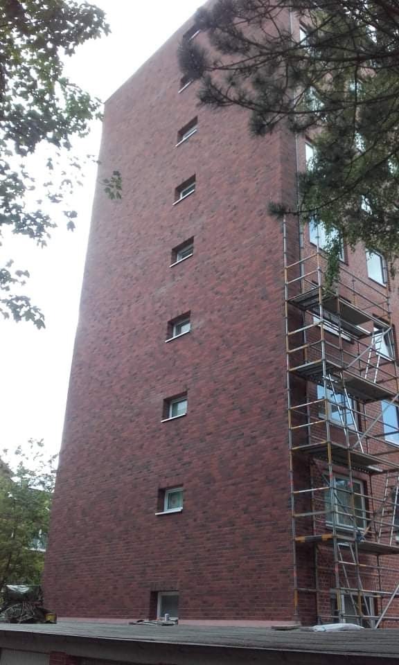 Wysoki budynek z czerwonej cegły z oknami, częściowo zakryty rusztowaniem, widok z dołu na tle drzew.