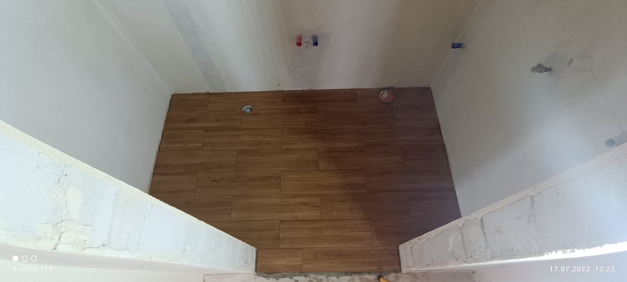 Ułożone brązowe płytki imitujące drewno na podłodze w surowym pomieszczeniu z widocznymi instalacjami i niedokończonymi ścianami.