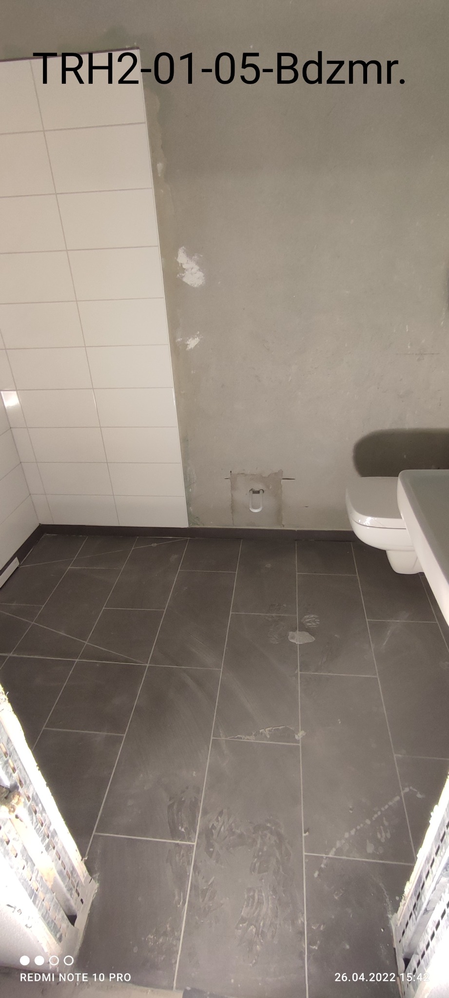 Wnętrze łazienki w trakcie remontu: ułożone ciemnoszare płytki podłogowe, białe płytki ścienne w rogu, surowa ściana z widocznymi śladami po pracach i częściowo zamontowana toaleta.