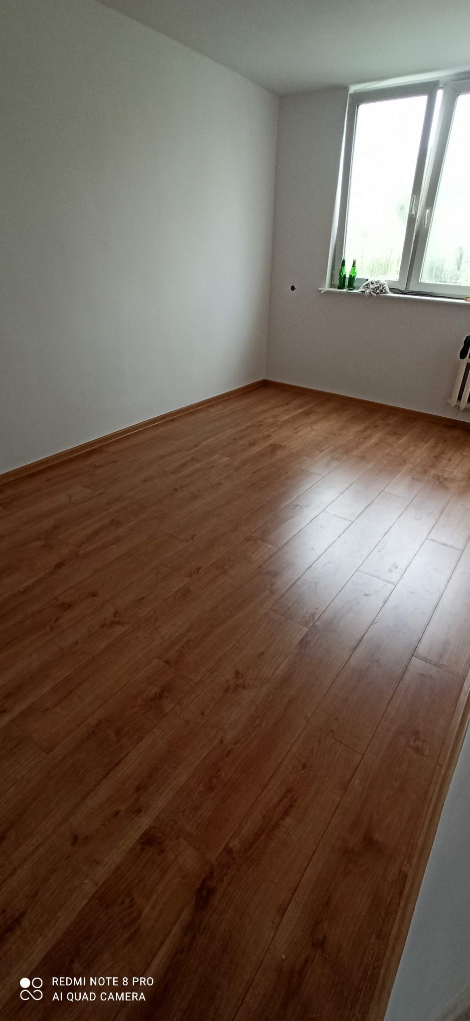 Pusty pokój z panelami podłogowymi w odcieniach brązu, świeżo pomalowane białe ściany, okno z butelkami i szmatką na parapecie.