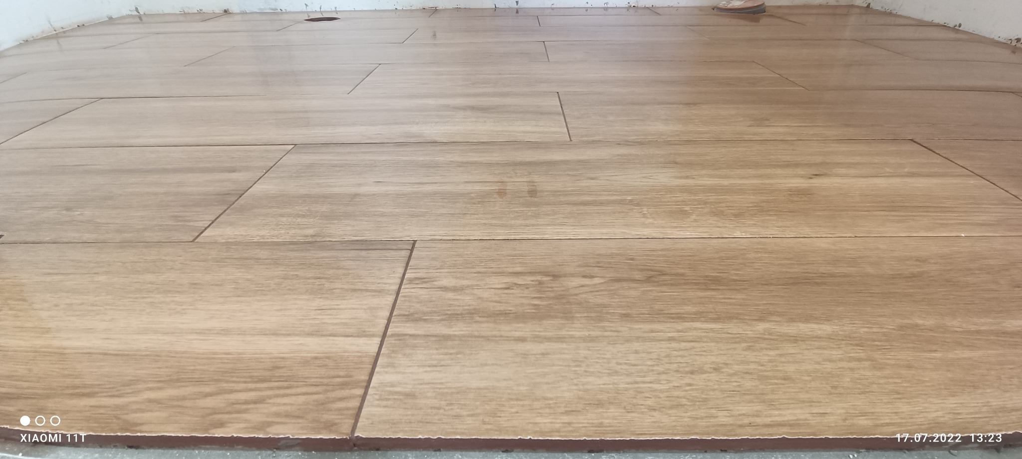 Ułożona podłoga z płytek imitujących drewno, widoczne fugi i pozostałości zaprawy przy ścianie.