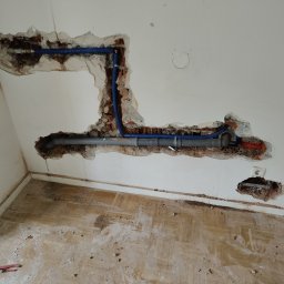 Phu Tomasz Sobieska - Ściana w trakcie prac instalacyjnych: widoczne niebieskie rury prowadzone w wykuciach, surowy stan, narzędzie na podłodze. Układanie instalacji wewnątrz budynku.