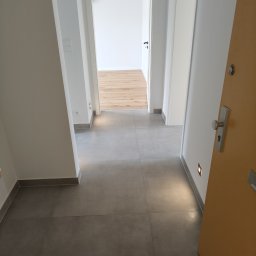 Phu Tomasz Sobieska - Nowoczesny korytarz z szarymi płytkami podłogowymi i minimalistycznym oświetleniem. W tle widoczne otwarte drzwi do pokoju z drewnianą podłogą.