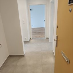 Phu Tomasz Sobieska - Jasne wnętrze mieszkania po wykończeniu: szare płytki podłogowe, białe ściany, widoczne drzwi wejściowe i otwarte drzwi do kolejnych pomieszczeń z drewnianą podłogą.