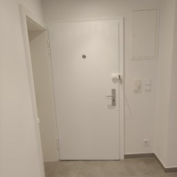 Phu Tomasz Sobieska - Białe drzwi wejściowe z wizjerem i zamkiem, widok na korytarz z płytkami podłogowymi. Świeże, minimalistyczne wykończenie wnętrza.