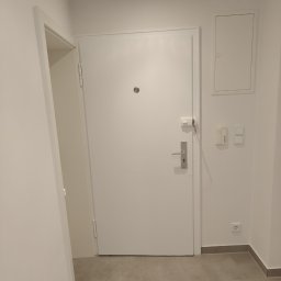 Phu Tomasz Sobieska - Białe drzwi wejściowe z wizjerem i srebrną klamką w jasnym korytarzu z szarą podłogą. Widoczny fragment otwartych drzwi po lewej i skrzynka elektryczna po prawej.