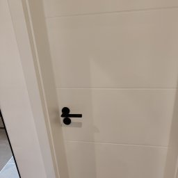 Phu Tomasz Sobieska - Białe drzwi z czarną klamką i subtelnymi poziomymi frezami, widoczne w kadrze od dołu, z fragmentem podłogi w tle.