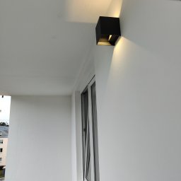 Phu Tomasz Sobieska - Biała ściana balkonu z kwadratową, czarną lampą ścienną, oświetlającą przestrzeń ciepłym światłem. Widoczne okno z białymi ramami i fragment budynku w tle.