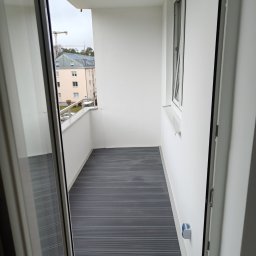 Phu Tomasz Sobieska - Nowoczesny, wykończony balkon z szarą podłogą z desek kompozytowych, białe ściany i widok na budynek mieszkalny zza balustrady. Widok z wnętrza przez otwarte drzwi balkonowe.