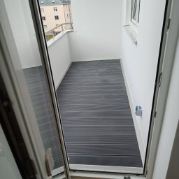 Phu Tomasz Sobieska - Nowoczesny balkon z szarą podłogą z desek kompozytowych, widok z wnętrza przez otwarte drzwi balkonowe, na zewnątrz budynek mieszkalny i dźwig budowlany.