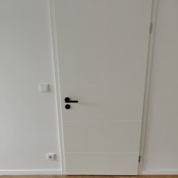 Phu Tomasz Sobieska - Nowoczesne, białe drzwi wewnętrzne z czarną klamką na tle białej ściany i drewnianej podłogi. Minimalistyczny design z poziomymi frezami.