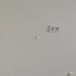 Gładka, biała ściana z zamontowanym podwójnym gniazdkiem elektrycznym i czujnikiem dymu, wykończona szarą listwą przypodłogową i płytkami podłogowymi.