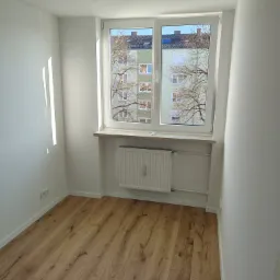 Jasny pokój po remoncie: drewniana podłoga, białe ściany, okno z widokiem na budynek. Grzejnik pod oknem. Minimalistyczny wystrój, czyste linie.