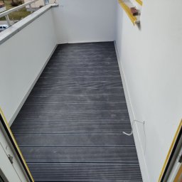 Phu Tomasz Sobieska - Wykończony balkon w Wolnzach z szarą podłogą z desek kompozytowych, białe ściany i balustrady. Widok z góry przez otwarte drzwi balkonowe.